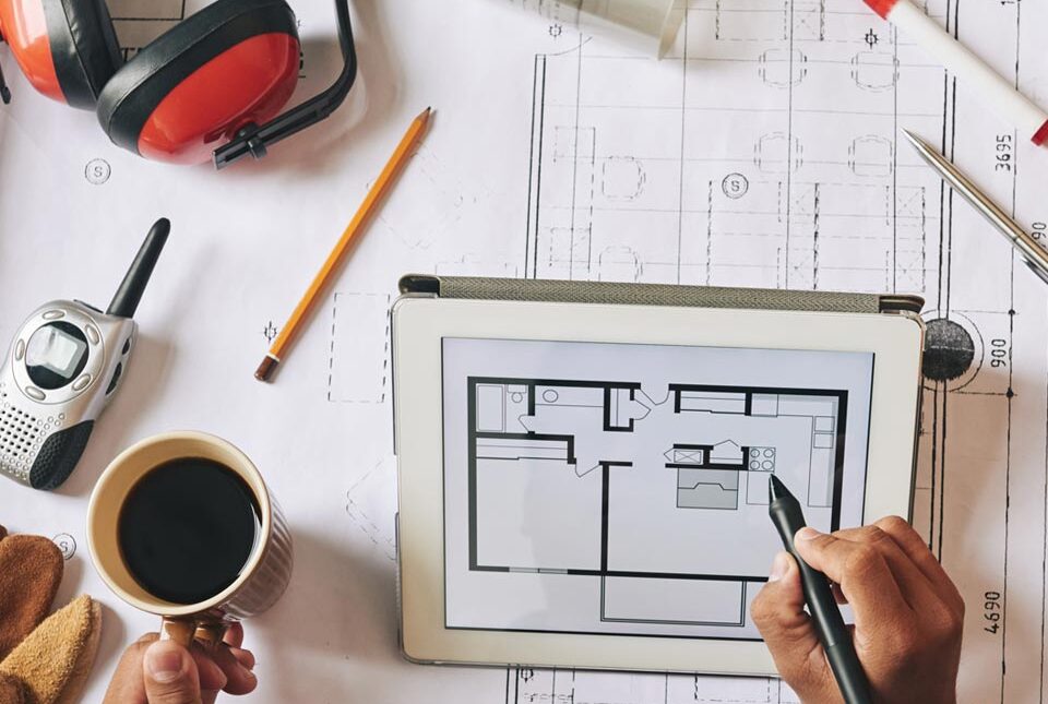 A contractor reviewing project blueprints on a tablet and at a drafting desk, surrounded by tools and equipment