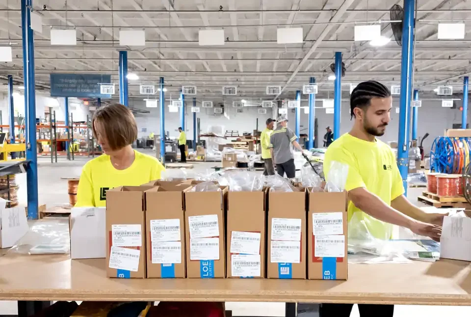 A pair of Loeb Electric warehouse associates prepare a kitting package to be shipped to an active jobsite. Six labeled cardboard boxes sit front and center.